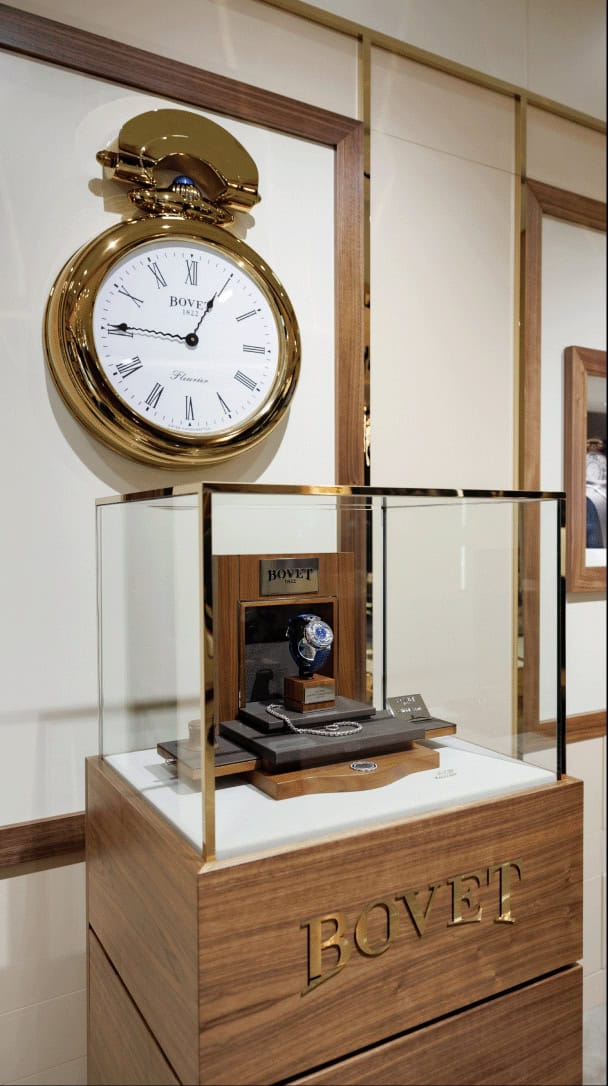 Elegant Bovet timepiece displayed in a glass case with a large vintage clock in the background.
