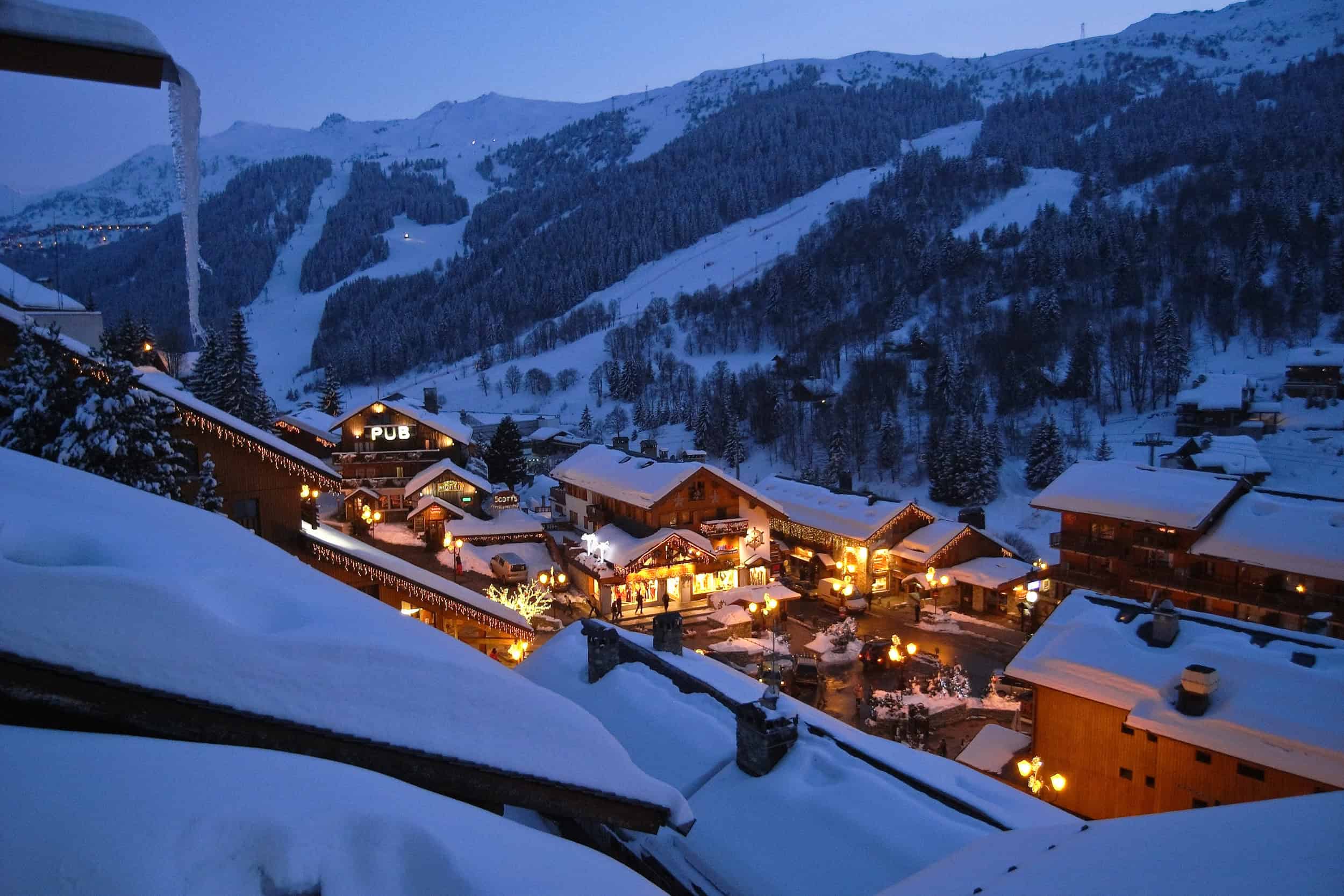 Snowy mountain village with illuminated chalets and ski slopes at dusk.