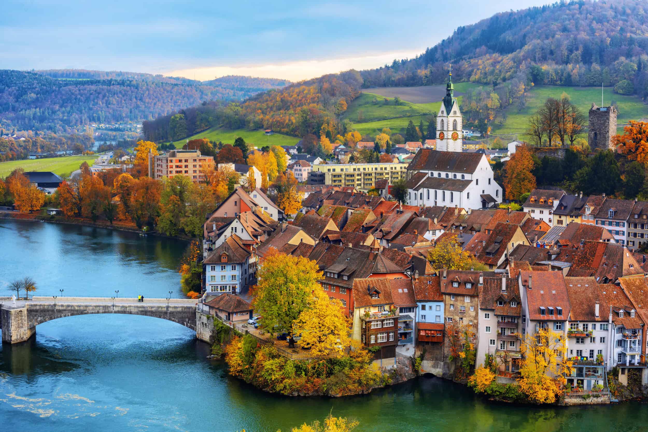 A colorful Swiss town with a river, bridge, and historic buildings surrounded by autumn foliage and.