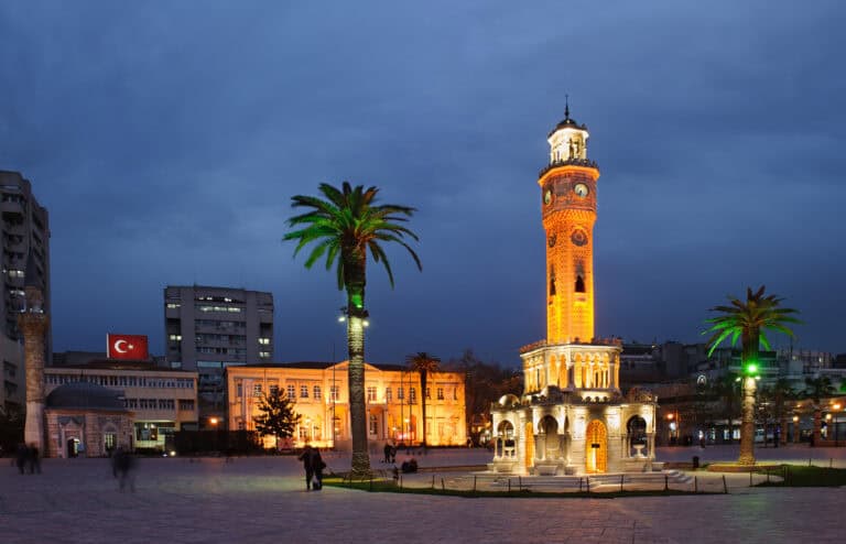 Iconic clock tower in Argos illuminated at dusk with palm trees and cityscape background.