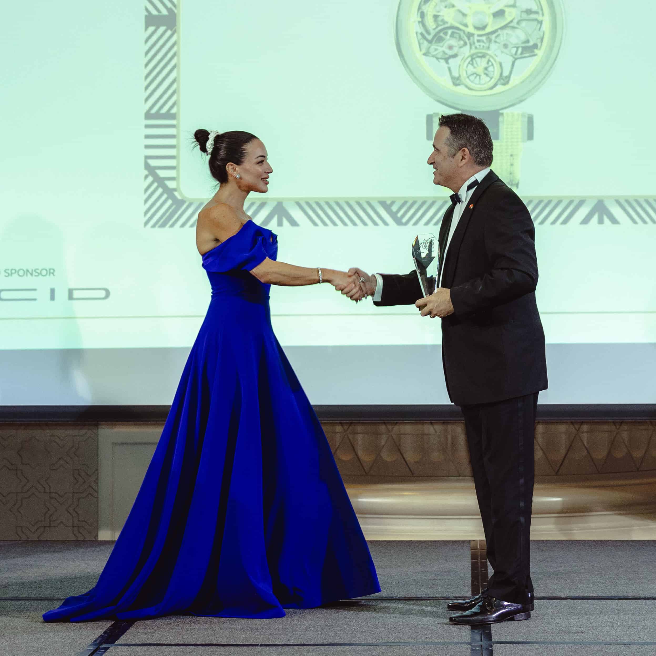 A stunning woman in a royal blue gown shaking hands with a man in a tuxedo during an award ceremony.